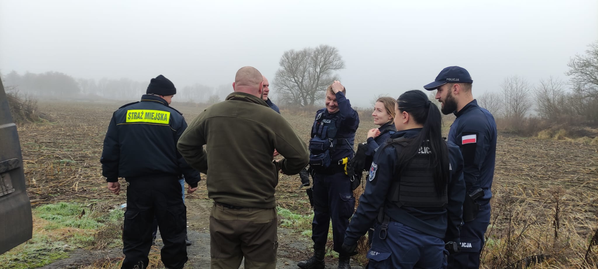 Strażnicy, policjanci oraz mężczyzna w zielonej bluzie i spodniach stoją na polu i rozmawiają ze sobą.