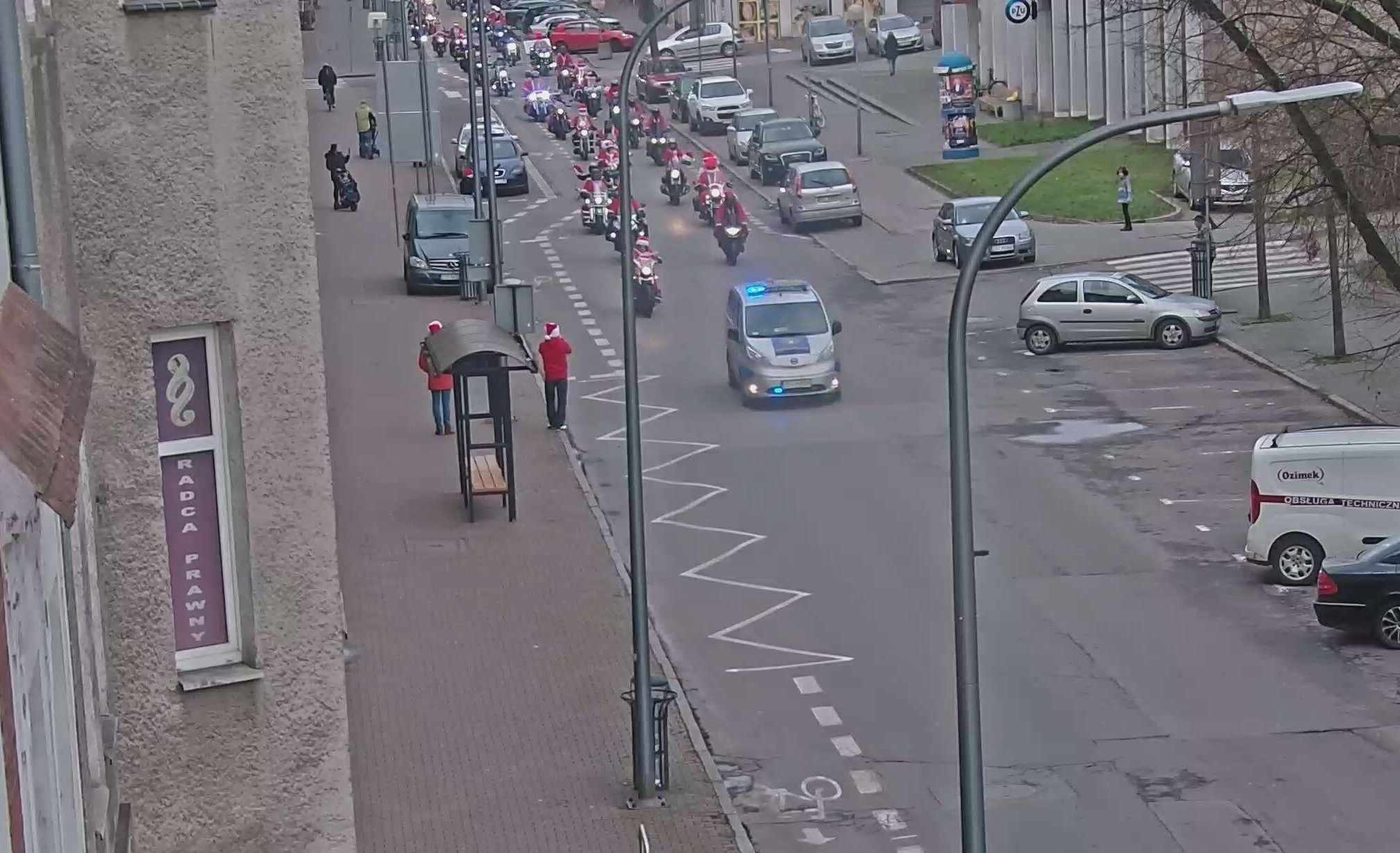 Ulica z szeroką jezdnią. Wzdłuż ulicy zaparkowane samochody. Na czele, środkiem jezdni, jedzie radiowóz Straży Miejskiej z włączonym kogutem. Dalej, w dwóch równoległych kolumnach, na kilkunastu motorach, jadą motocykliści ubrani w strój Świętego Mikołaja. Idący chodnikiem przechodnie przyglądają się wydarzeniu.