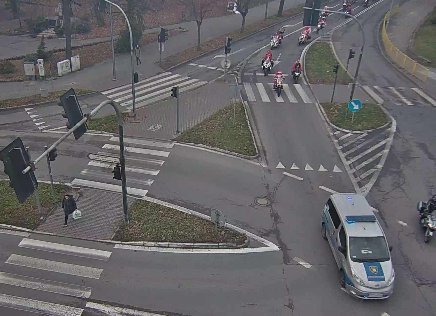 Przez skrzyżowanie przejeżdża grupa kilkunastu motocyklistów ubranych w stroje Świętego Mikołaja. Na czele grupy jedzie radiowóz Straży Miejskiej z włączonym kogutem. Motocykliści jadą za radiowozem jeden za drugim w dwóch równoległych kolumnach.