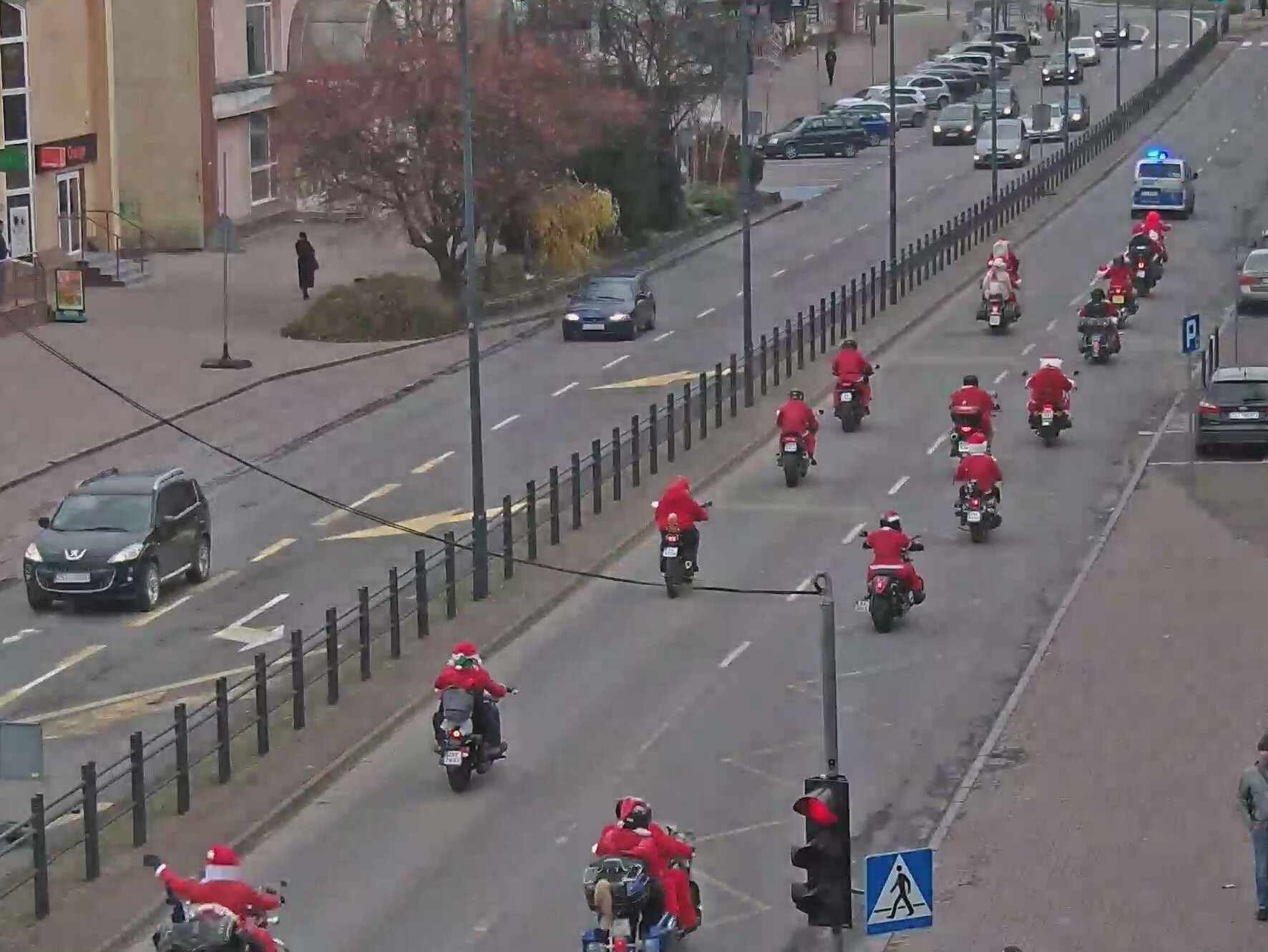 Kilkunastu motocyklistów ubranych w stroje Świętego Mikołaja, jedzie całą szerokością dwujezdniowej drogi prowadzeni przez radiowóz Straży Miejskiej. Motocykliści Jadą jeden za drugim w dwóch równoległych kolumnach.