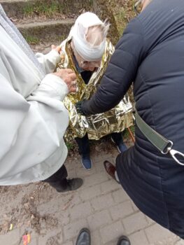 Na chodniku siedzi starszy mężczyzna z opatrunkiem na głowie i owinięty kocem ratunkowym. Obok stoją udzielające pomocy osoby.
