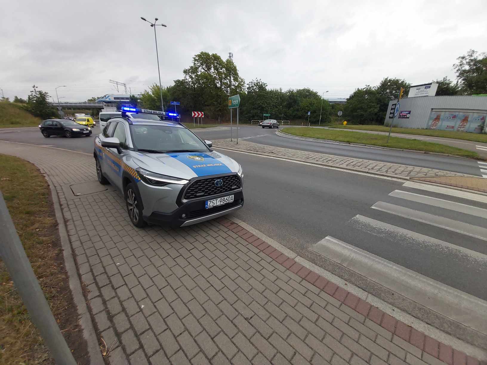 Radiowóz Straży Miejskiej, który stoi na chodniku z włączonymi sygnałami świetlnymi przy rondzie.