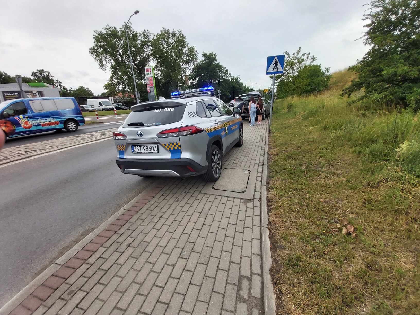 Radiowóz Straży Miejskiej, który stoi na chodniku z włączonymi sygnałami świetlnymi. Dalej również na chodniku stoi uszkodzony samochód osobowy.