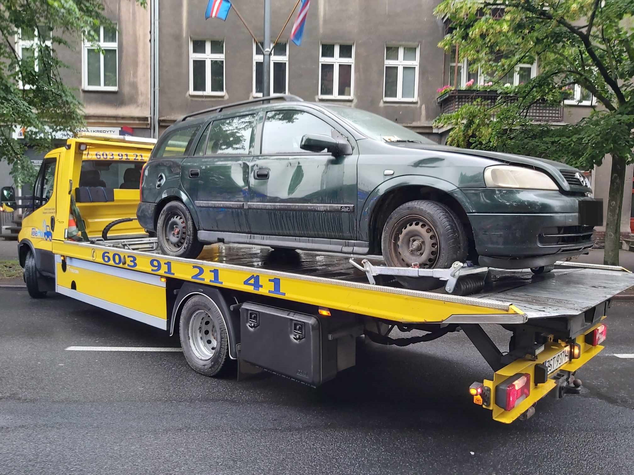 Na ulicy manewruje laweta z załadowanym uszkodzonym zielonym samochodem marki opel.