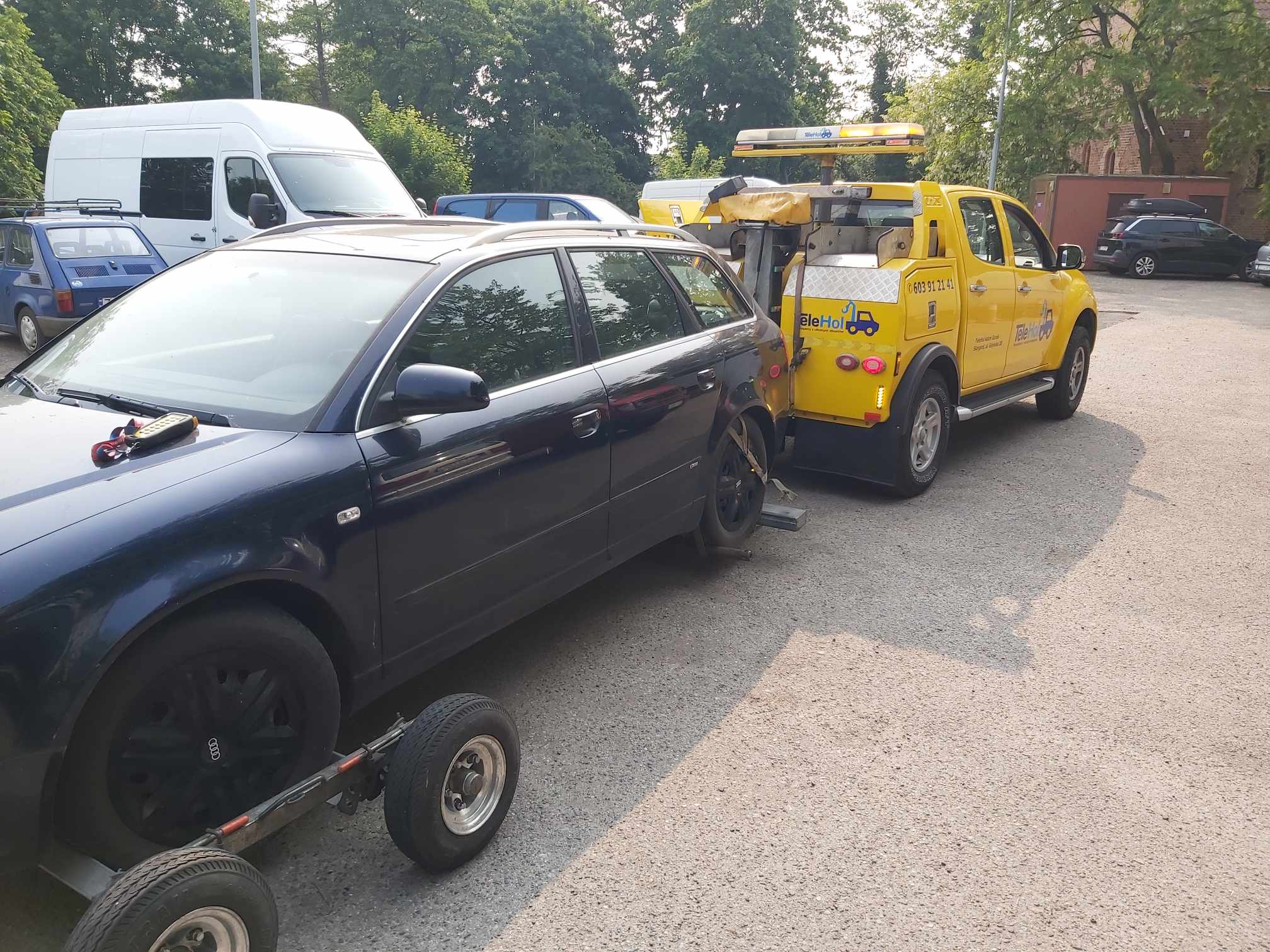 Przy parkingu stoi holownik z przyczepionym uszkodzonym samochodem marki audi.