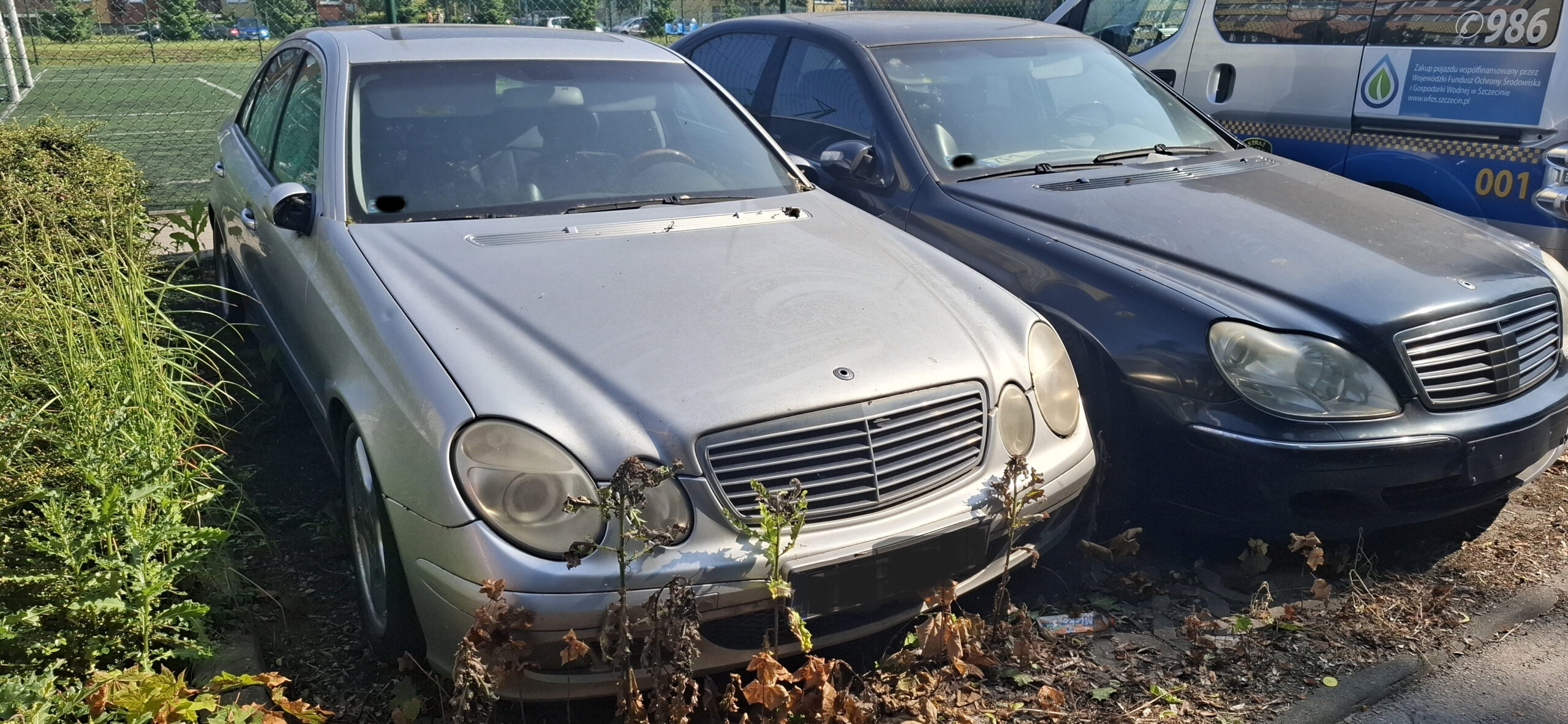 Na parkingu stoją obok siebie dwa zaniedbane i uszkodzone samochody marki mercedes.