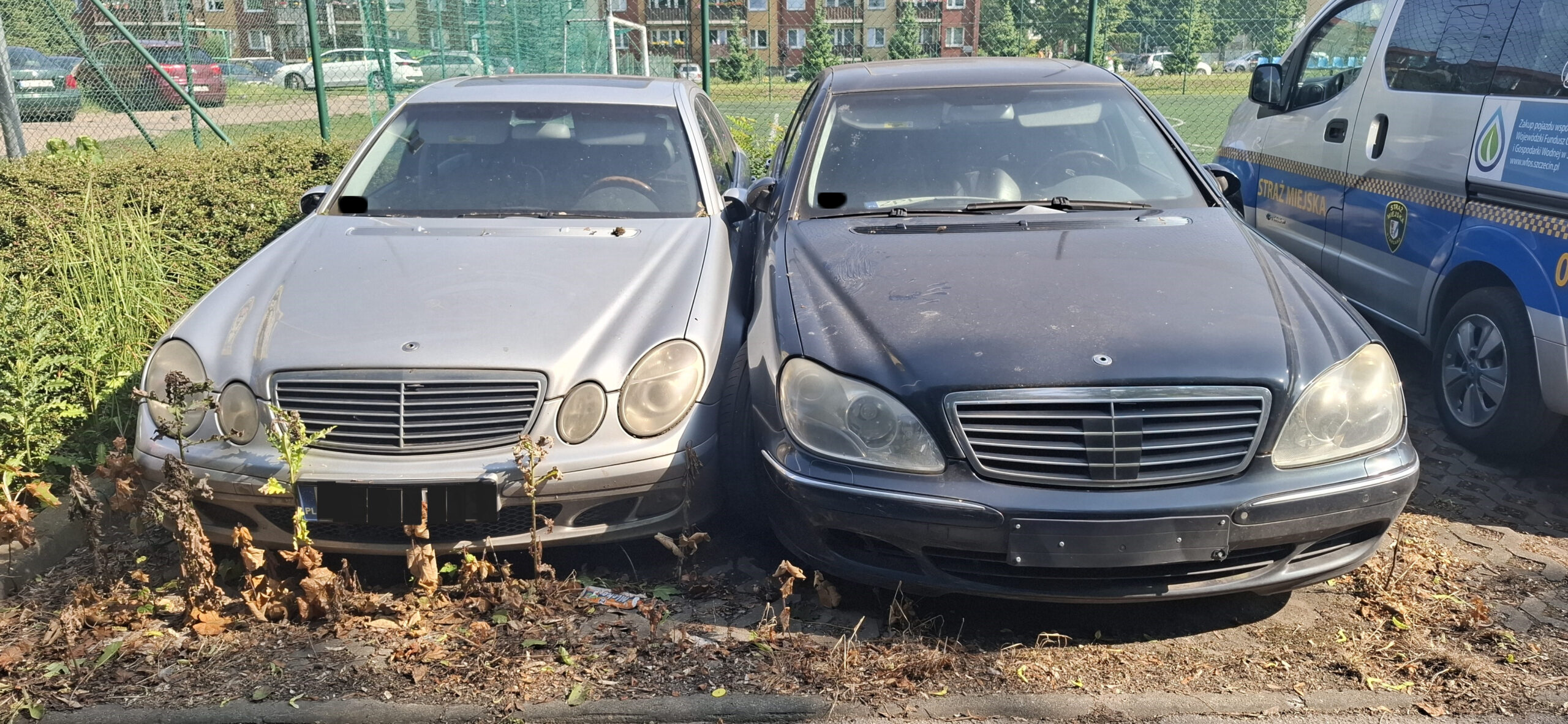 Na parkingu stoją obok siebie dwa zaniedbane i uszkodzone samochody marki mercedes.