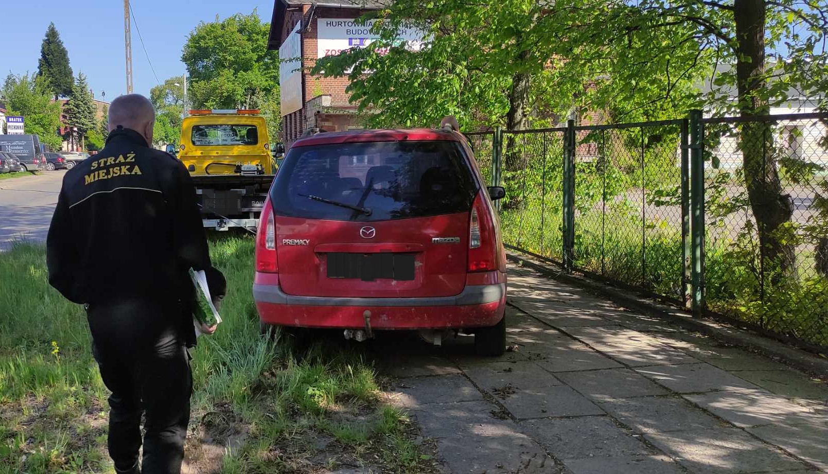 Laweta wciąga zdezelowany samochód marki mazda, który stał na trawniku i chodniku. Z tyłu stoi strażnik miejski.