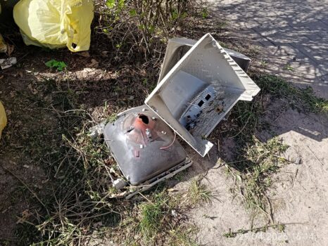 Rozbity kineskop telewizora i plastikowa obudowa porzucone na ziemi obok chodnika, w pobliżu krzewów i worka na odpady.