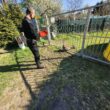 Mężczyzna w rękawicach ochronnych stoi przy metalowej bramie i obserwuje dwa psy znajdujące się po drugiej stronie ogrodzenia.